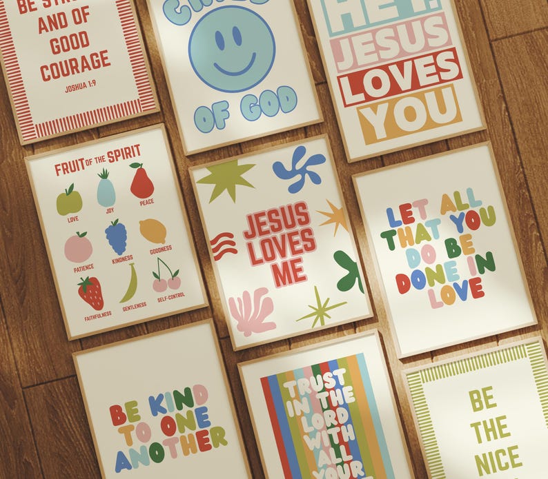 May include: A collection of framed art prints with inspirational quotes and colorful designs. The prints feature phrases like "Jesus Loves Me," "Be Kind to One Another," and "Fruit of the Spirit." The artwork is displayed on a wooden surface.