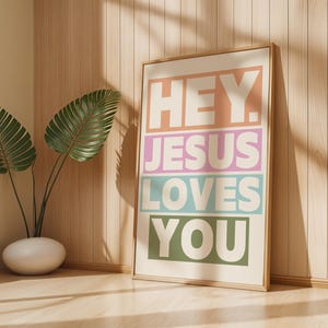 May include: A framed art print with the words "HEY. JESUS LOVES YOU" in a colorful, block-letter design. The print is in a light wood frame and leans against a light wood wall. A potted plant with large green leaves is in the corner.