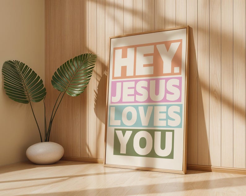 May include: A framed art print with the words "HEY. JESUS LOVES YOU" in a colorful, block-letter design. The print is in a light wood frame and leans against a light wood wall. A potted plant with large green leaves is in the corner.