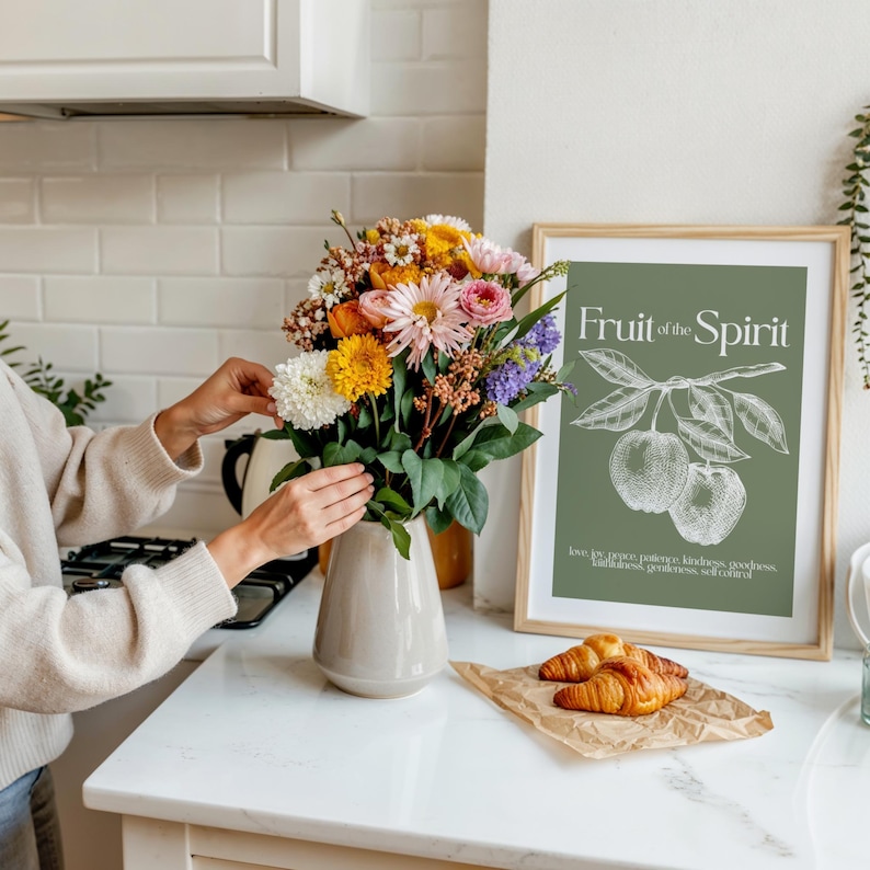 May include: A colorful bouquet of flowers in a beige vase, with a framed print that reads "Fruit of the Spirit." Croissants rest on a piece of brown paper. The scene is set in a kitchen with white cabinets and a white countertop.
