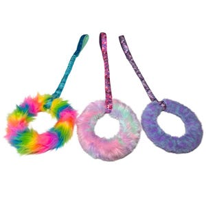 Könnte beinhalten: Drei kreisförmige Tierspielzeuge mit Ringen aus flauschigem Kunstfell, die an Stoffbändern befestigt sind. Der erste Ring ist regenbogenfarben, der zweite pastellfarben in Rosa und Blau und der dritte in Violett und Blau gehalten.