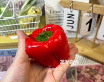 Könnte beinhalten: Ein leuchtend roter Paprika mit einem grünen Blattdetail wird in einer Hand gehalten. Der Paprika ist glatt und glänzend. Der Hintergrund umfasst einen Kalender mit dem 1. Juni und ein Blumengesteck.
