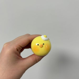May include: A small yellow ceramic chick figurine with a white egg on its head. The chick has black eyes and an orange beak.