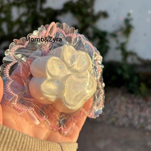 Puede incluir: Un derrite de cera blanco en forma de flor con acentos de purpurina iridiscente, empaquetado en celofán transparente con un ribete de encaje rosa y blanco. El texto "Momo&Zyra" es visible.