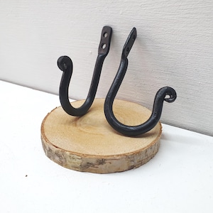 Könnte beinhalten: Zwei schwarze Metallhaken, die auf einem Holzsockel montiert sind. Die Haken sind S-förmig und haben ein rustikales, handgeschmiedetes Aussehen.