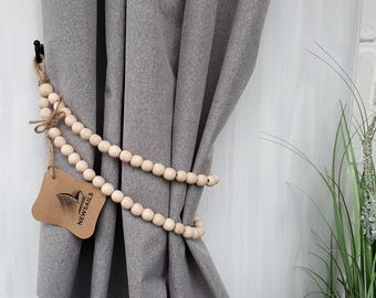 May include: A set of wooden beads in a light brown color are used as a curtain tieback. The beads are wrapped around a gray curtain and tied with a brown string. A small brown tag with the text "NEWSAILS" is attached to the beads.