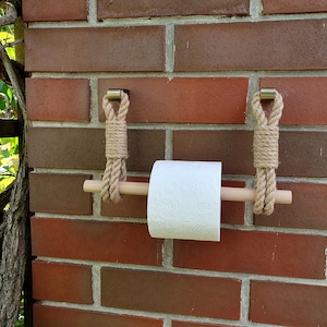 May include: A rustic toilet paper holder made of natural rope and a wooden dowel. The holder is suspended from a brick wall with metal brackets. A roll of white toilet paper is in place, ready for use.
