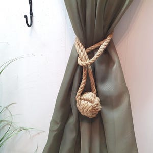 May include: A brown rope curtain tieback with a knotted ball at the end. The tieback is holding back a green curtain.