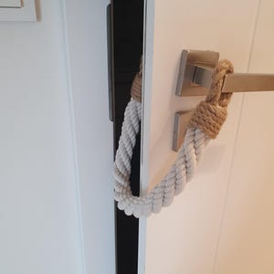 May include: A white twisted rope door stopper featuring tan jute rope accents at both ends. The rope is looped around a brushed metal door handle and extends to the other side of the door, keeping it slightly ajar against a white door frame.