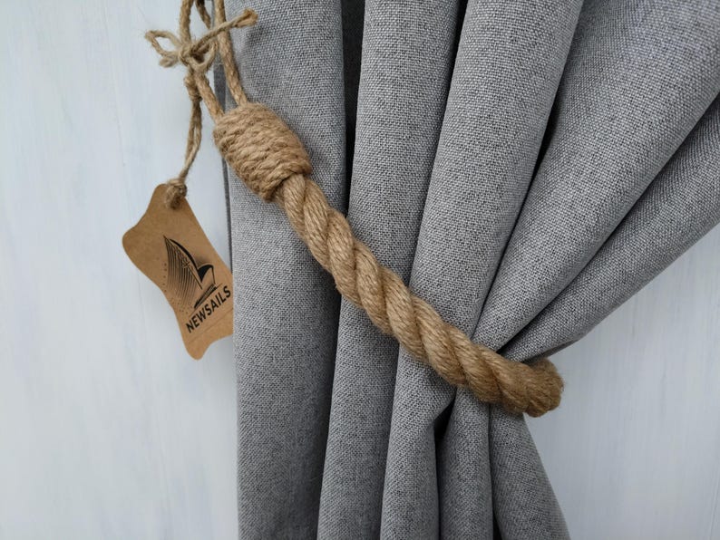 Pode incluir: Um amarrador de cortina de corda natural grossa em um tecido cinza claro com uma etiqueta marrom que diz "NEWSAILS".