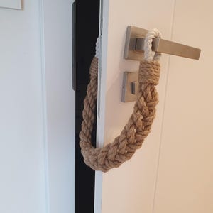 May include: A door stopper made of thick, braided tan jute rope. The rope is looped over a brushed metal door handle, with a white cord section at the top. The braided rope hangs down in a U-shape, providing a physical barrier to keep the door open.