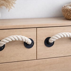 Puede incluir: Primer plano de una cómoda de madera clara con dos cajones. Cada cajón tiene un tirador de cuerda sujeto a una placa de metal negro. Los tiradores son de cuerda gruesa de color blanquecino. La cómoda está en una habitación con decoración neutra.