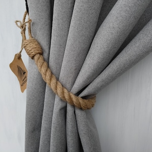 Pode incluir: Cortinas cinzas amarradas com um amarrador de corda natural grosso. O amarrador &eacute; feito de uma corda natural grossa e tem um n&oacute; simples na ponta. O amarrador &eacute; amarrado em torno das cortinas e mantido no lugar por um gancho de metal.