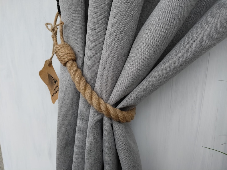 Pode incluir: Cortinas cinzas amarradas com um amarrador de corda natural grosso. O amarrador &eacute; feito de uma corda natural grossa e tem um n&oacute; simples na ponta. O amarrador &eacute; amarrado em torno das cortinas e mantido no lugar por um gancho de metal.