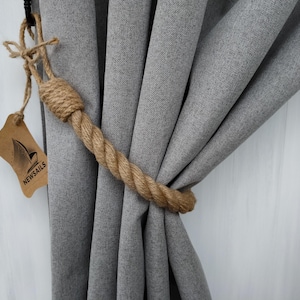 Pode incluir: Close-up de uma cortina cinza com um amarrador de corda marrom. O amarrador &eacute; feito de corda natural e tem um design de n&oacute; simples. A etiqueta anexada ao amarrador diz "NEWSAILS".