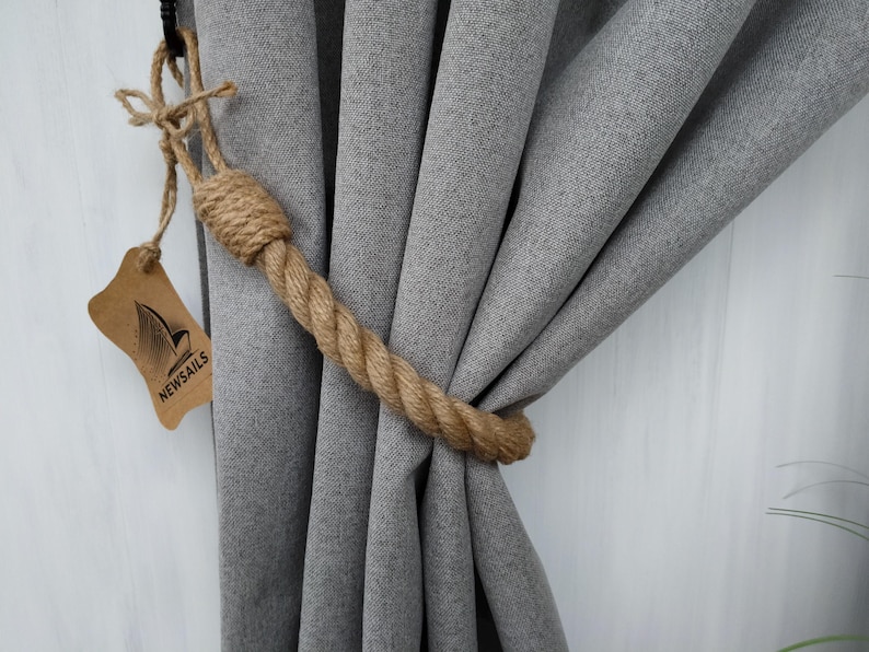 Pode incluir: Close-up de uma cortina cinza com um amarrador de corda marrom. O amarrador &eacute; feito de corda natural e tem um design de n&oacute; simples. A etiqueta anexada ao amarrador diz "NEWSAILS".