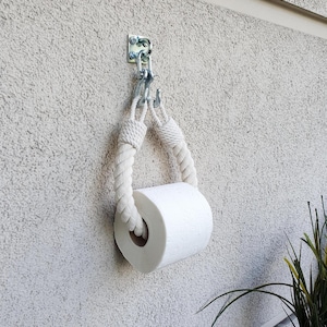 May include: A wall-mounted toilet paper holder. The holder is made of a thick, off-white rope suspended from a metal bracket. A roll of white toilet paper is in the holder. The holder is attached to a textured, light-colored wall.