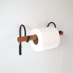 May include: A black metal and wood toilet paper holder mounted on a white wall. The holder features a wooden dowel for the toilet paper roll and a black metal bracket.