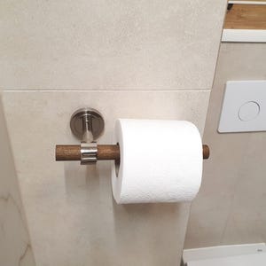 May include: A modern toilet paper holder with a wooden dowel and a brushed nickel mounting plate. The holder is mounted on a light beige tiled wall, with a roll of white toilet paper in place.