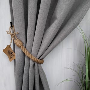 Pode incluir: Cortina cinza com um amarrador de corda. O amarrador &eacute; feito de corda de juta natural e est&aacute; preso a um gancho de metal preto. O amarrador de corda est&aacute; etiquetado com "NEWSAILS".