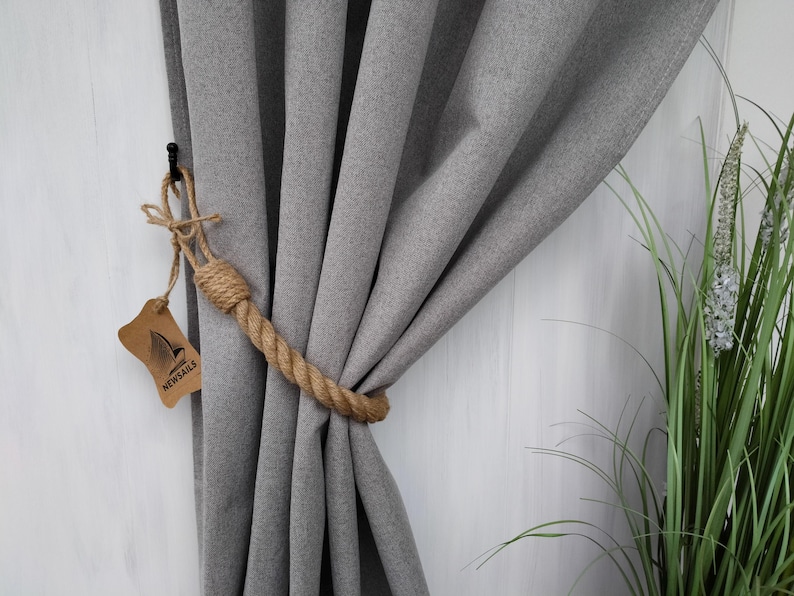Pode incluir: Cortina cinza com um amarrador de corda. O amarrador &eacute; feito de corda de juta natural e est&aacute; preso a um gancho de metal preto. O amarrador de corda est&aacute; etiquetado com "NEWSAILS".