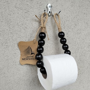 May include: A wooden bead toilet paper holder with a natural jute rope and black wooden beads. The holder is hanging from a hook on a wall. The text "NEWSAILS" is printed on a brown tag.
