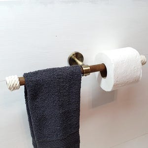 May include: A rustic bathroom fixture featuring a wooden towel bar with rope accents and a matching toilet paper holder. A dark gray towel hangs below the bar, and a roll of white toilet paper is on the holder. The fixture is mounted on a white wall.