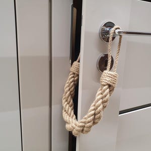 Puede incluir: Un tirador de puerta decorativo hecho de gruesa cuerda beige. La cuerda está anudada en la parte superior e inferior, formando un lazo que cuelga del tirador de la puerta. La puerta es blanca con un tirador plateado.