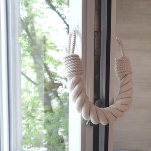 Puede incluir: Un tirador de puerta de cuerda blanca con un lazo en el extremo. El tirador está sujeto a un marco de ventana blanco.
