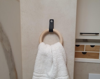 May include: A white towel hangs from a wooden ring towel holder with a black leather strap. The holder is mounted on a light beige wall. The towel is folded and has a textured appearance.