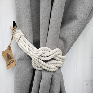 Può includere: Ferma tendina in corda bianca con un nodo al centro. Il ferma tendina è attaccato a una tenda grigia. L'etichetta sul ferma tendina recita "New Sails".