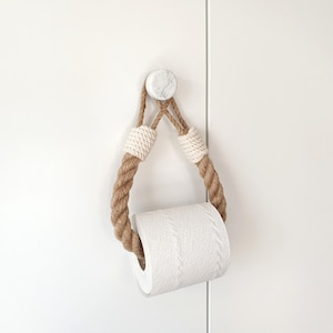 May include: A rustic toilet paper holder made of thick, twisted brown rope, with white woven accents. The holder is suspended from a marble-look knob and holds a roll of white toilet paper. The design is simple and nautical.