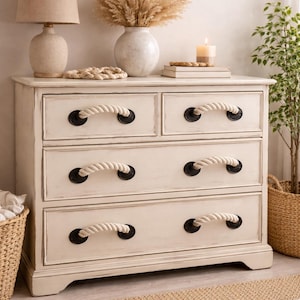 May include: Cream-coloured wooden chest of drawers with four drawers, each with rope handles and black hardware. The dresser is part of a coastal or nautical-themed bedroom set. A lamp, vase, and mirror are visible.
