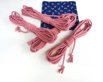 May include: Several coils of pink rope with knotted ends are arranged on a white surface. A navy blue fabric with a white anchor pattern is in the background. The ropes are thick and appear to be made of cotton.