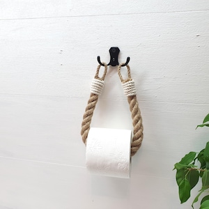 May include: A rustic toilet paper holder made from natural rope with white accents. The holder is attached to a black metal hook and hangs on a white wall. A roll of toilet paper is on the holder.