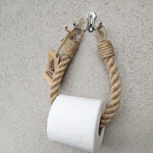 May include: A nautical-themed toilet paper holder made of thick, beige rope. The rope is looped and attached to a silver hook, holding a roll of white toilet paper. A small tag with a sailboat logo hangs from the rope.