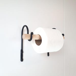 May include: A black metal toilet paper holder with a wooden dowel supporting a roll of white toilet paper. The holder is mounted on a white wall, showcasing a minimalist design.