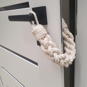 May include: A white door with a black handle and a decorative rope door stop. The door stop is made of thick, braided rope and is looped over the handle. The door is white with horizontal grooves.