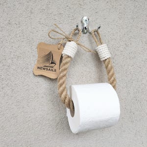 May include: A white toilet paper roll holder made of natural rope with white rope ends. The holder is attached to a wall with a silver hook. The holder has a brown tag with the text "NEWSAILS" and a sailboat graphic.