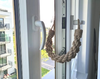 May include: A window with a white frame and a white handle. A decorative rope handle is attached to the window frame. The rope is a light brown color and is looped to form a handle. The window is open, and a view of the outside is visible.