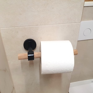 May include: A black metal and wood toilet paper holder mounted on a beige tiled wall. The holder features a black circular base, a wooden dowel, and a roll of white toilet paper. The design is minimalist and modern.