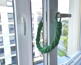 May include: A green braided rope window handle. The handle is looped and attached to a white window frame. The window is open, revealing a view of a building and trees.
