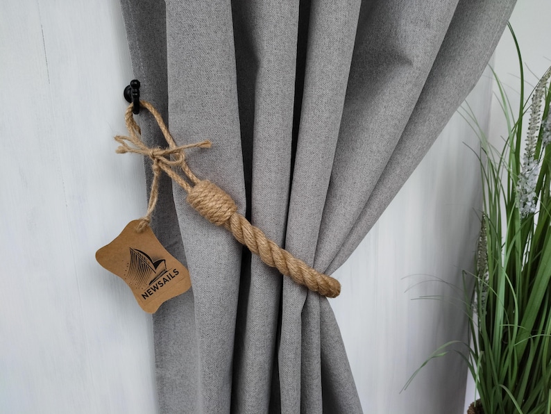 Pode incluir: Close-up de uma cortina cinza com um amarrador de corda. O amarrador &eacute; feito de corda de juta natural e est&aacute; amarrado em um n&oacute;. Uma pequena etiqueta marrom com o texto "NEWSAILS" est&aacute; presa &agrave; corda.