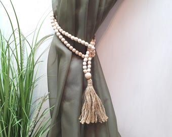 May include: A set of wooden beads with a jute tassel, used as a curtain tieback. The beads are natural in color and the tassel is a light brown. The tieback is wrapped around a green curtain.