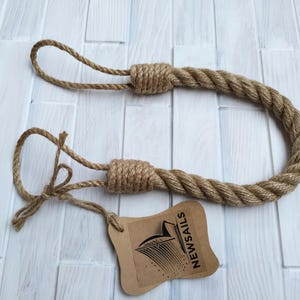 Pode incluir: Um amarrador de cortina de corda marrom com duas al&ccedil;as nas pontas. O amarrador &eacute; feito de corda de fibra natural e tem um design simples e r&uacute;stico. O amarrador &eacute; etiquetado com uma etiqueta marrom que diz "NEWSAILS".
