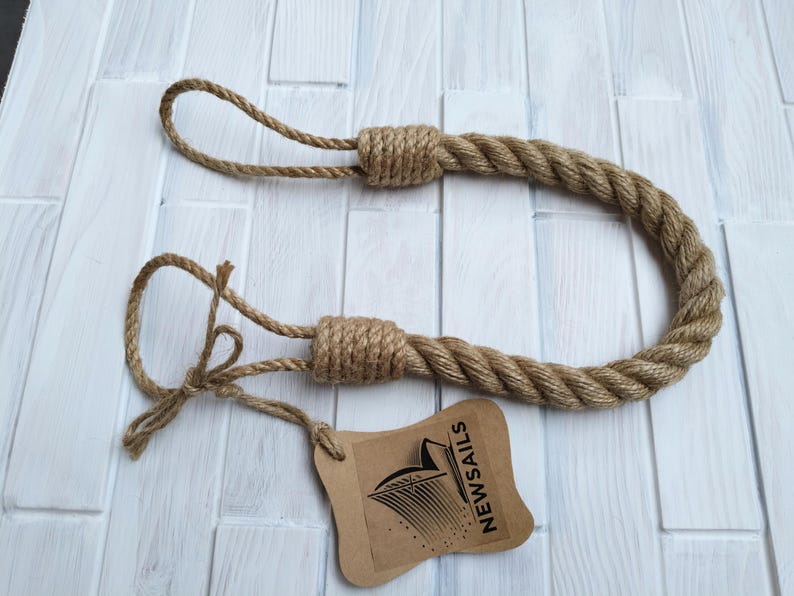 Pode incluir: Um amarrador de cortina de corda marrom com duas al&ccedil;as nas pontas. O amarrador &eacute; feito de corda de fibra natural e tem um design simples e r&uacute;stico. O amarrador &eacute; etiquetado com uma etiqueta marrom que diz "NEWSAILS".
