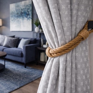 May include: A light grey curtain secured with a brown rope tieback and a black square fitting. The rope is looped around the curtain, adding a decorative touch. The background reveals a living room with a blue sofa and abstract artwork.