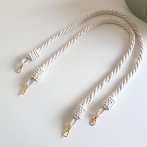 May include: Cream-coloured rope bag straps with gold-tone clasps. The straps are made of twisted rope and feature decorative knots. The bag straps are shown on a white surface, with a portion of a green bag visible in the background.