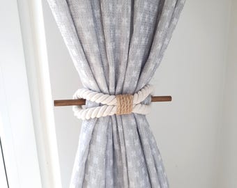 May include: A curtain tieback made of thick, off-white rope and a brown wooden dowel. The rope is wrapped around the curtain and secured with a natural jute twine. The curtain is a light gray with a subtle white pattern.