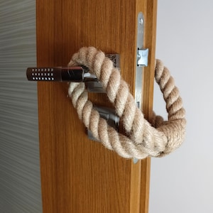 Pode incluir: Um close-up de uma maçaneta de porta com uma corda enrolada ao redor. A corda é de cor marrom claro e está amarrada em um nó. A maçaneta da porta é feita de metal e tem um acabamento prateado.